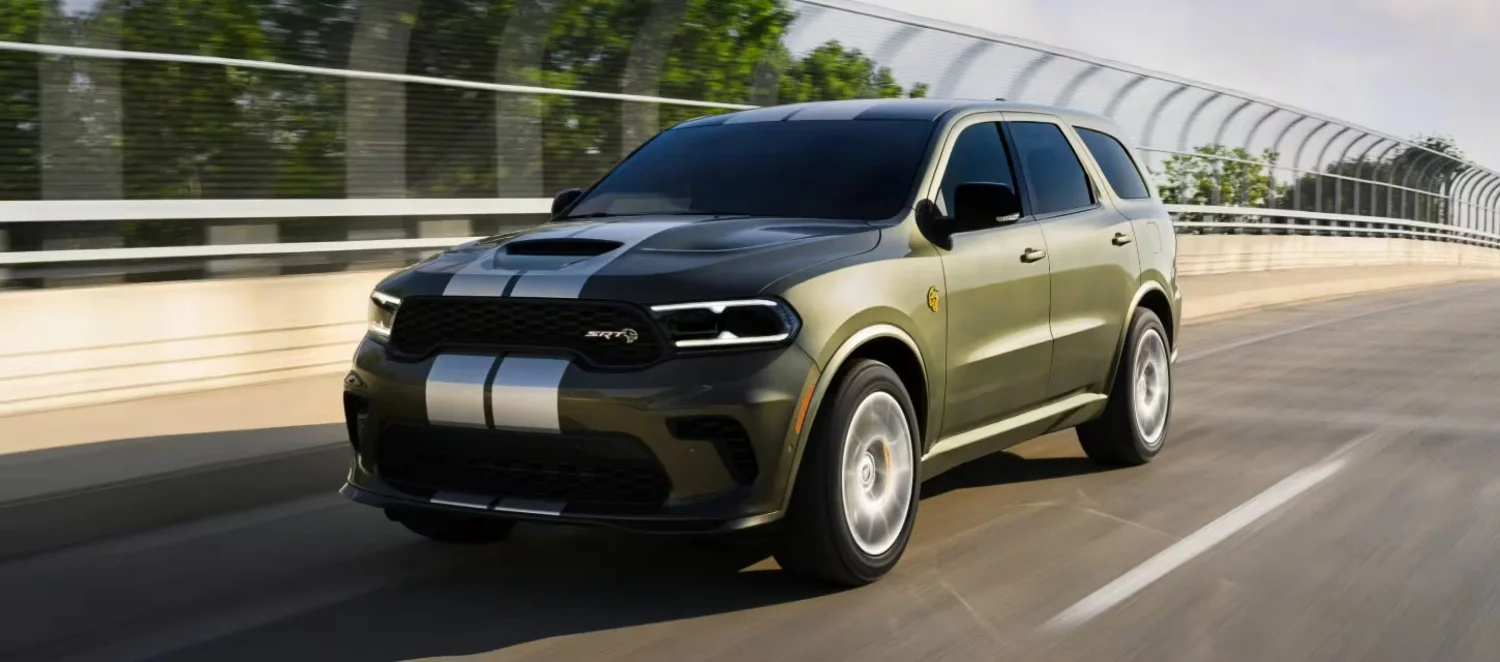 Koyu haki rengindeki 2027 Dodge Durango performans SUV modeli, kaputunda ve ön tamponunda uzanan gümüş renkli çift şeridi ve SRT logosuyla, hareket halinde olduğu asfalt bir yolda sağa doğru hafifçe dönmüş şekilde güçlü görünümünü sergiliyor.