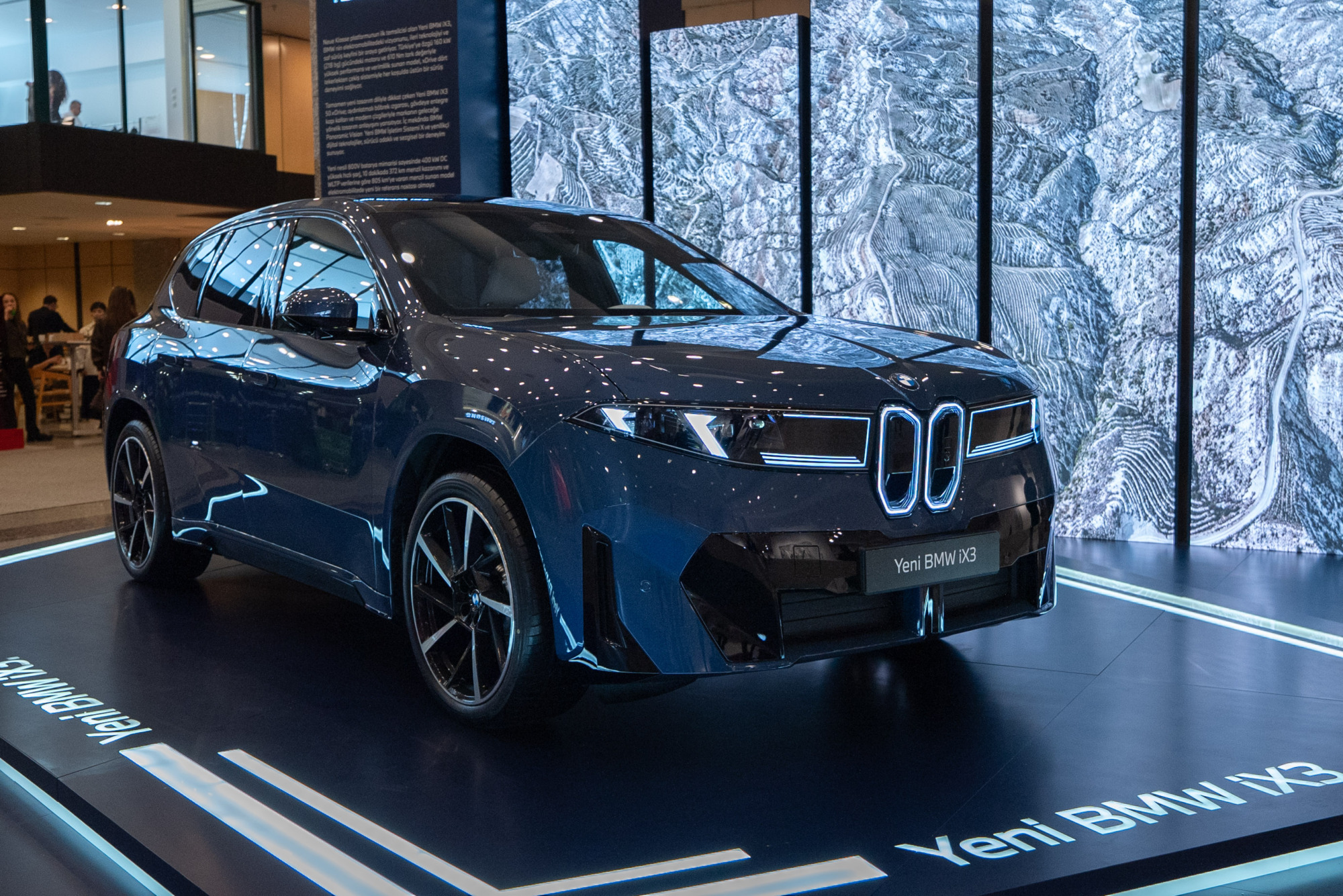 Koyu mavi renkli, Yeni BMW iX3 elektrikli SUV, modern aydınlatmalı ön ızgarası ve farlarıyla parlak siyah bir teşhir platformunda göz alıcı bir şekilde sergilenirken, arka plandaki dağlık araziyi yansıtan büyük ekranlar teknolojik bir atmosfer oluşturuyor.