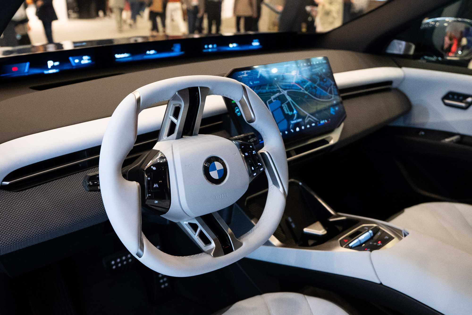 Beyaz direksiyon simidi, üzerinde belirgin bir BMW logosu ve entegre kumandaları olan modern bir otomobil kokpiti, yatay dijital gösterge paneli ve geniş navigasyon haritası gösteren merkezi bilgi-eğlence ekranıyla birlikte yüksek teknoloji ve çağdaş bir iç tasarım sunuyor.