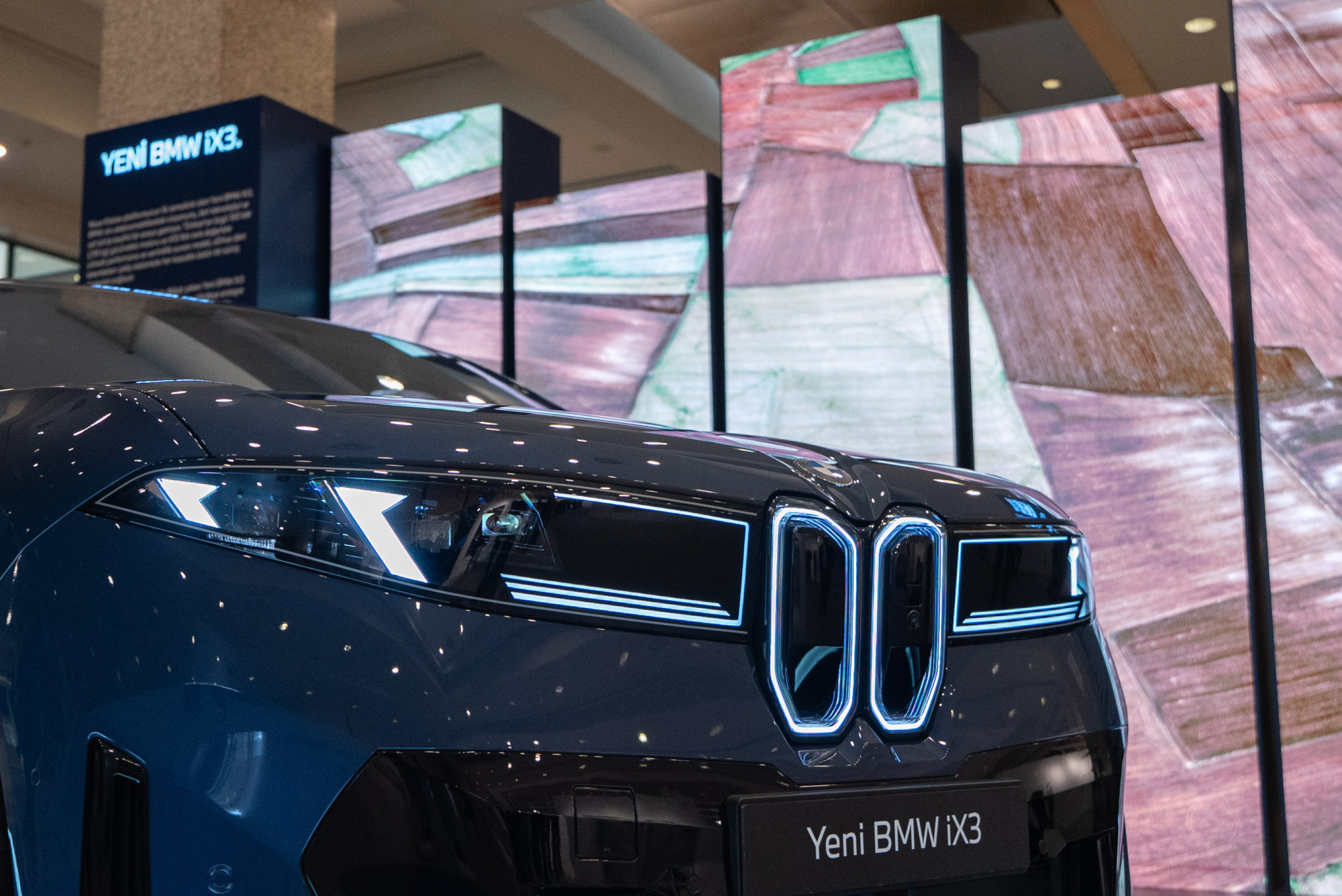 Karanlık bir ortamda parlak farları ve belirgin mavi ışıklı ızgarasıyla dikkat çeken Yeni BMW iX3'ün ön kısmından detaylı bir görüntü; arka planda ise soyut renkli dijital ekranlar ve model adının yazdığı panolar görülebiliyor.
