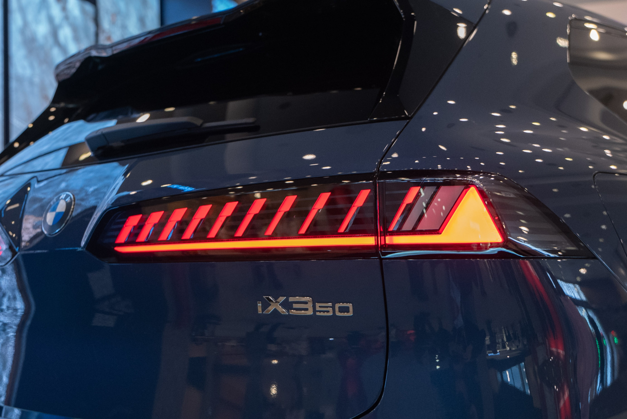 Koyu mavi renkli modern bir BMW iX3 50 elektrikli aracın arka sol köşesi yakın çekim olarak gösterilmiştir; aracın parlak yüzeyinde tavan ışıklarının yansımaları belirginken, yanan kırmızı dikey LED segmentleri ve altında uzun turuncu bir şerit bulunan stop lambası dikkat çekmektedir.