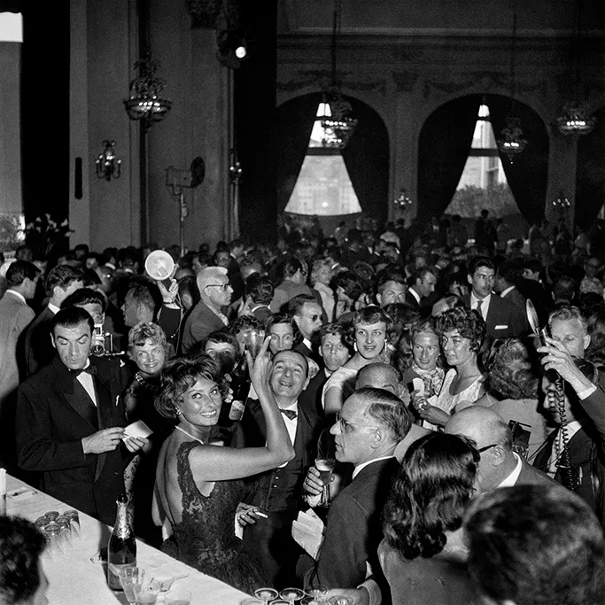Ara Güler'in 1959 Cannes sergisi fotoğrafında, zarif siyah elbisesiyle kalabalık bir partide neşeyle gülümseyen Sophia Loren, çevresindeki konuklarla birlikte canlı bir anın merkezinde, bar masasının yanında objektife poz veriyor.