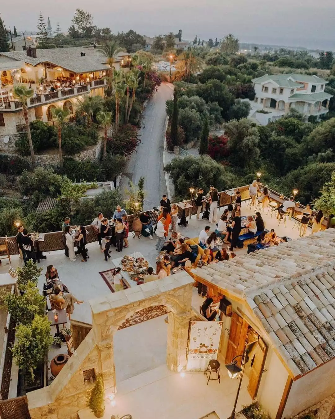 Alacakaranlıkta yukarıdan çekilmiş bu fotoğrafta, taş kemerli bir avlunun ötesinde, aydınlatılmış binaların ve yemyeşil ağaçların ortasında kıvrılan bir yolun görüldüğü bir manzaraya sahip, geniş bir açık hava terasında sohbet eden, yemek yiyen ve içeceklerin tadını çıkaran kalabalık bir grup insan betimleniyor.