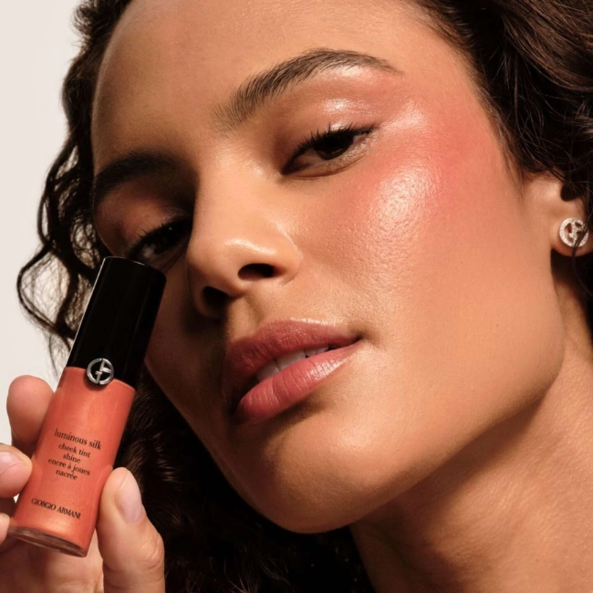 Cildinde ipeksi ve ışıltılı bir dokunuşla belirginleşen yanakları ve göz kapaklarıyla, elinde Giorgio Armani Luminous Silk Cheek Tint Shine ürününü tutan bir modelin çarpıcı yakın çekim portresi.