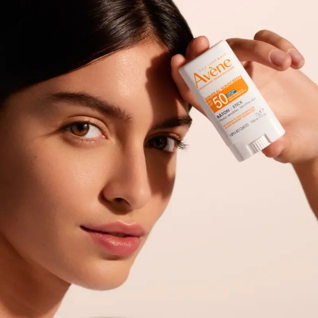 Esmer saçlı ve kahverengi gözlü genç bir kadın, hassas ciltler için özel olarak formüle edilmiş Avene Mineral Güneş Koruyucu SPF 50 Çubuk ürününü yüzünün önünde tutarak, güneşin zararlı etkilerinden korunmanın önemini vurguluyor.