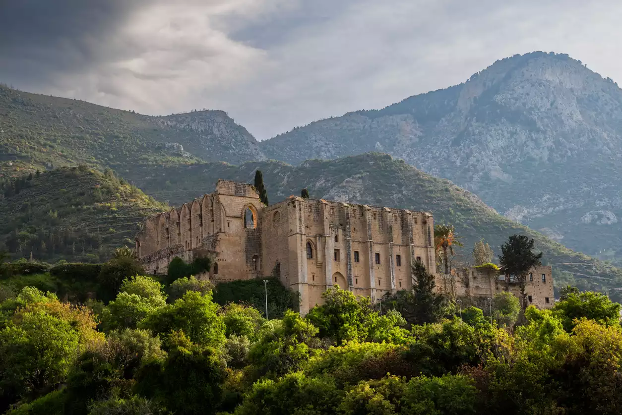 Bulutlu gökyüzü altında, yemyeşil ağaçların ortasında yükselen, kemerli pencereleri ve sağlam taş duvarlarıyla Bellapais Manastırı'nın etkileyici görüntüsü, arkasında sıralanan heybetli dağlarla birlikte tarihi ve doğal güzelliği bir araya getiriyor.