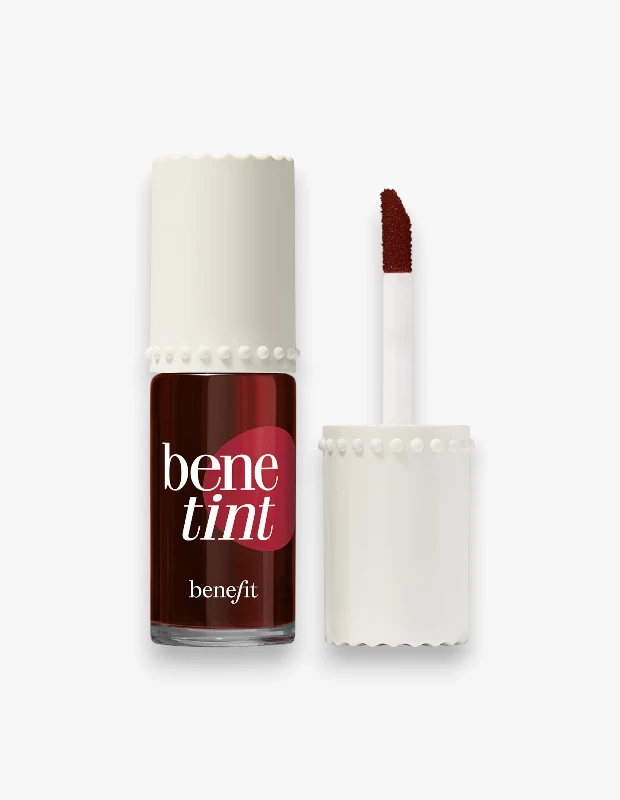 Beyaz düz zeminde konumlandırılmış, şeffaf cam şişesi içindeki koyu gül rengi Benefit Cosmetics Benetint dudak ve yanak renklendiricisinin, dalgalı kenarlı beyaz kapağı açık duruyor ve zengin rengini yansıtan aplikatörü yanında görülüyor.