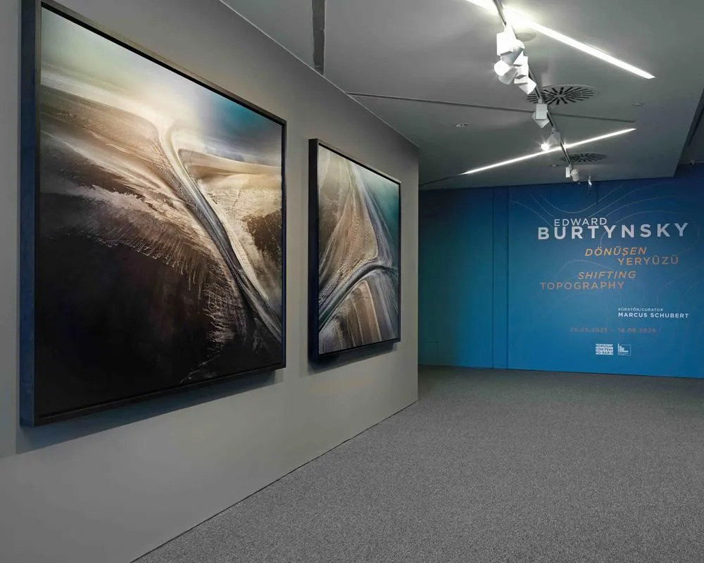 Borusan Contemporary'de Edward Burtynsky'nin Dönüşen Yeryüzü sergisinden, karanlık kahverengi ve açık mavi tonlarındaki iki büyük, havadan çekilmiş gibi görünen coğrafi formları yansıtan eserler gri duvarda sergileniyor; sağda ise serginin adı ve detayları mavi bir panelde belirgin şekilde yer alıyor.