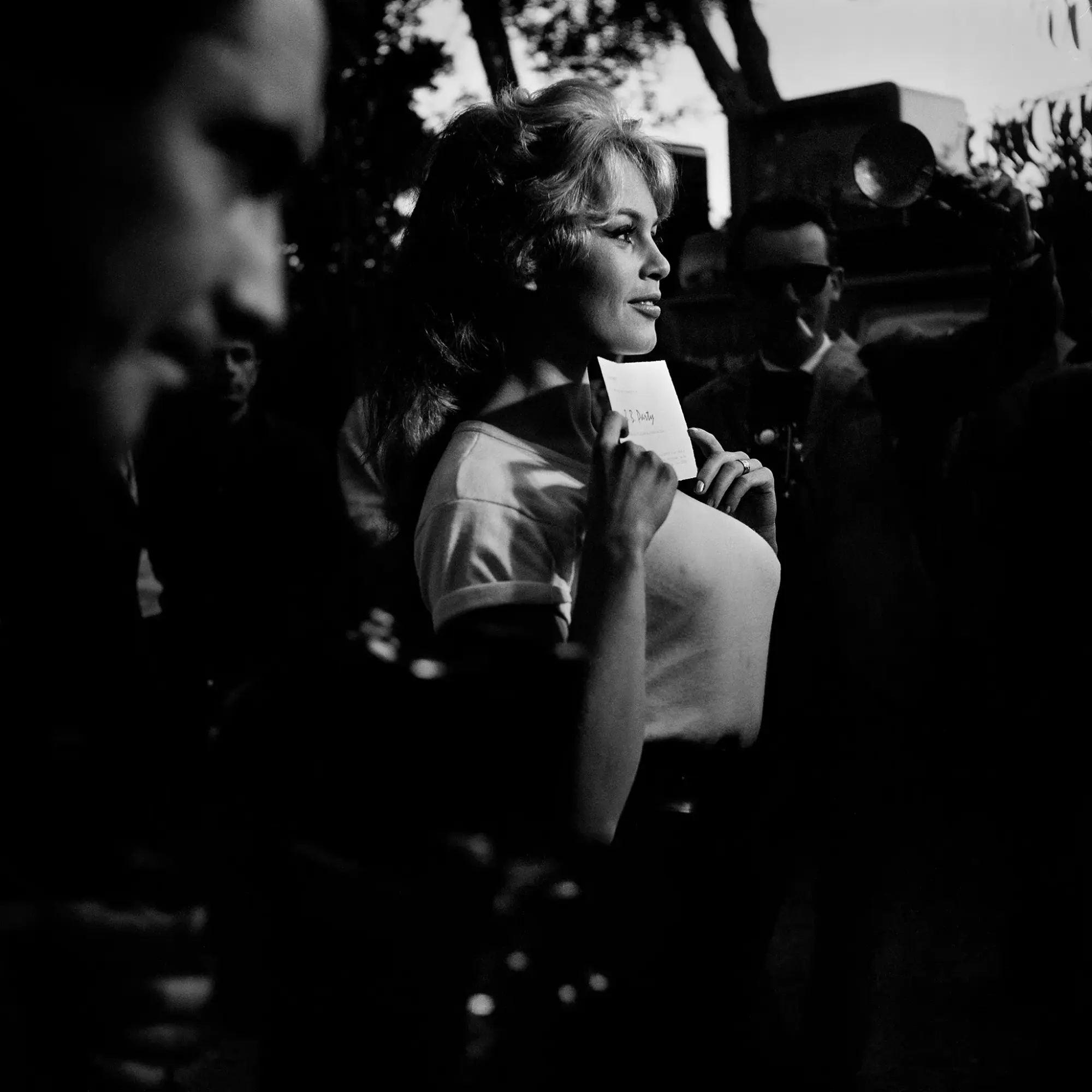 Siyah beyaz bu karede, 1957'den Brigitte Bardot, ikonik havalı sarı saçları ve zarif gülümsemesiyle profilinden sağa bakıyor, göğsüne doğru hafifçe tuttuğu üzerinde muhtemelen B. Bardot yazan bir kartı gösterirken, yüzü ve saçları kalabalık ve ağaçların oluşturduğu karanlık arka planda parlıyor.