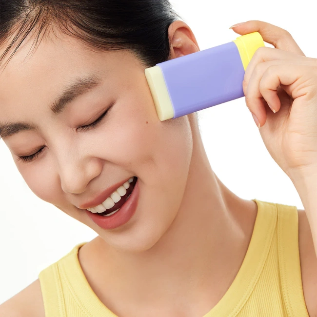 Mutlu bir gülümsemeyle gözleri kapalı, sarı atlet giyen genç bir kadın, mor ve sarı renkli By Wishtrend Bakuchiol Güneş Kremi Stiği'ni yanağına sürerken, bu SPF 50 korumalı ürünün keyifli ve kolay uygulanışını sergiliyor.