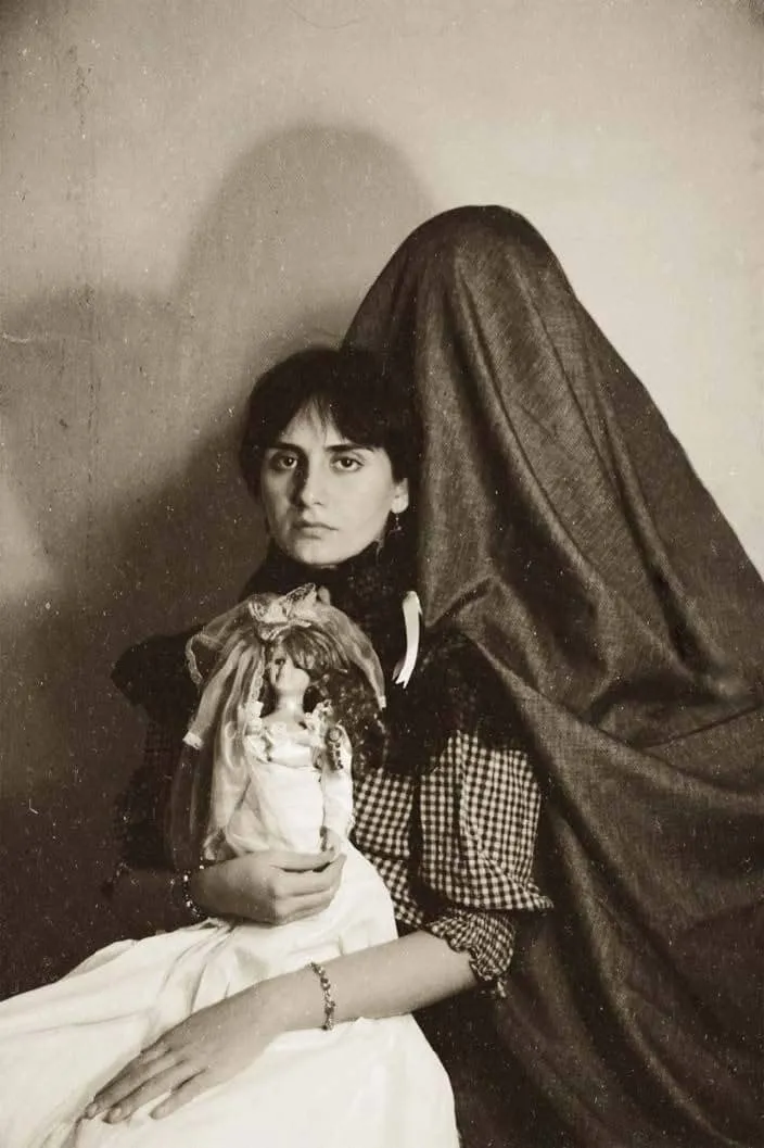 Selin Gencoglu'nun dijital fotoğrafında, ciddi bakışlı bir kadın kucağında gelinlikli bir oyuncak bebek tutarken, arkasında gizemli, örtülü bir figür adeta tekinsiz bir yokluk hissi yaratıyor.