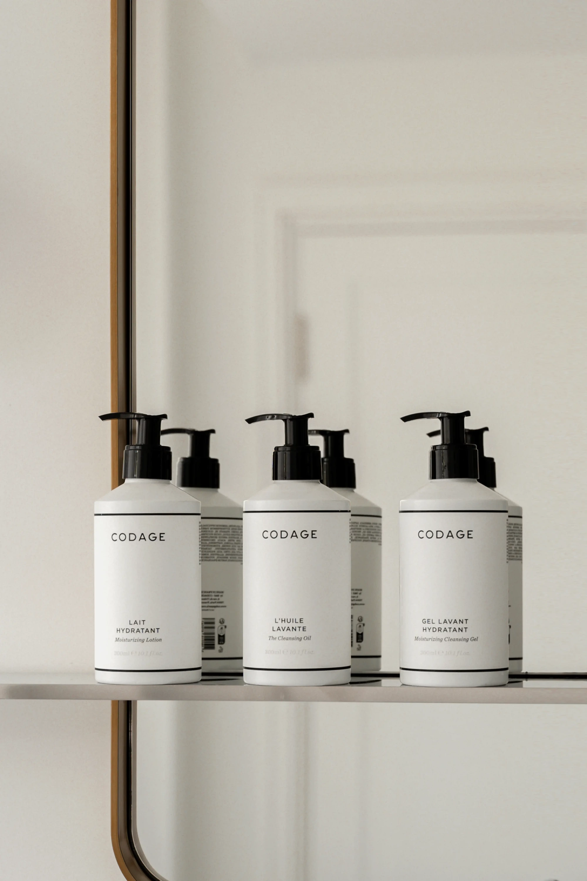 Bir rafta, her biri 200 ml hacimli, siyah pompa başlıklı ve CODAGE markalı, minimalist beyaz şişeler sıralanmış; ön plandakiler Nemlendirici Losyon, Temizleme Yağı ve Nemlendirici Temizleme Jeli olarak etiketlenmiş, arkada ise benzer şişelerin yansımasıyla modern ve lüks bir cilt bakım koleksiyonu sergileniyor.
