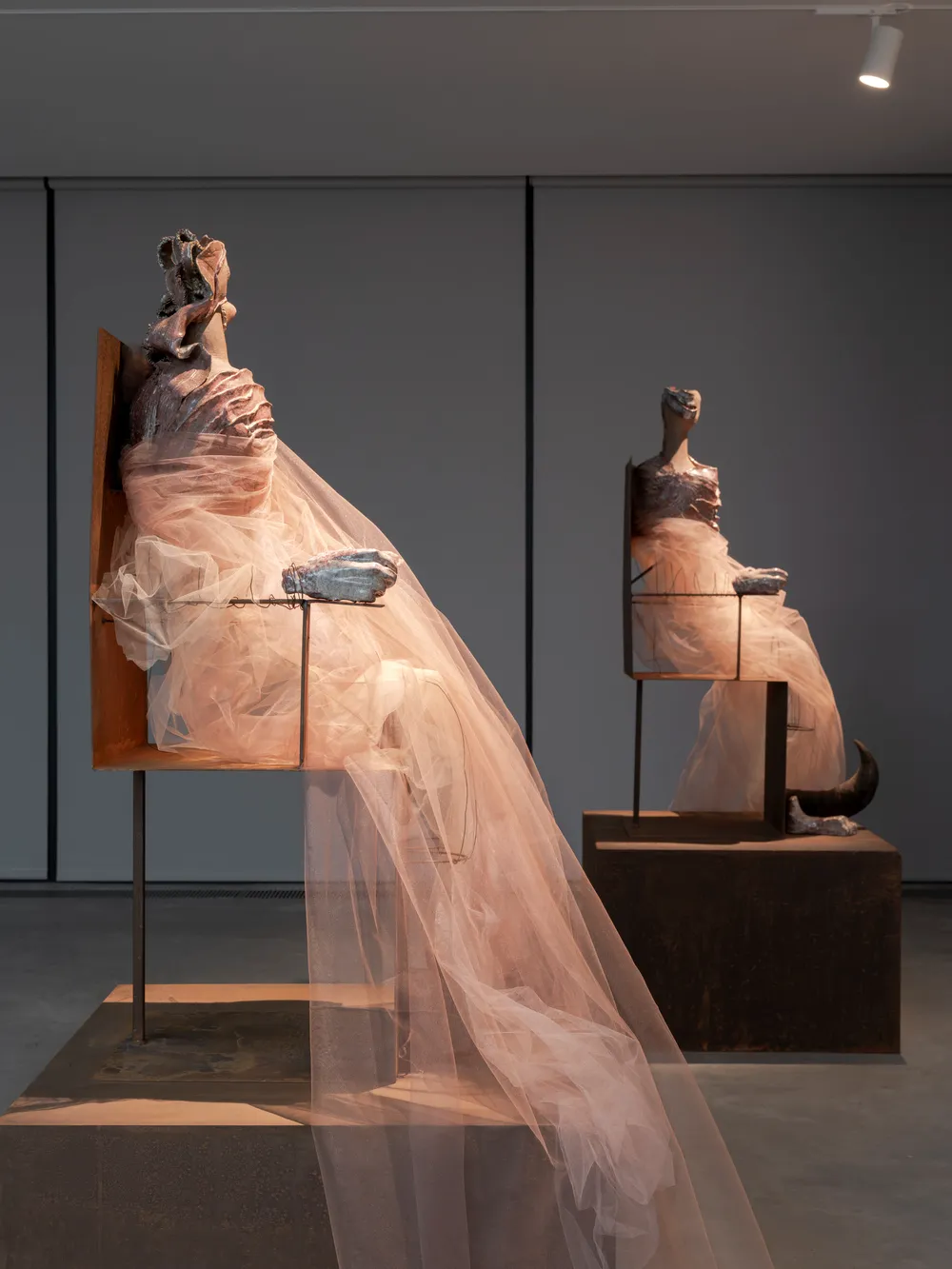 Canavarların Vaatleri temalı sergiye uygun olarak, ışıklandırılmış bir sanat galerisinde, pudra rengi yarı şeffaf tülden kumaşlarla gizemli bir şekilde örtülmüş iki soyut oturan figür heykeli, özellikle ön plandaki metal eliyle belirginleşen canavar figürünün sunulan vaatleri üzerine düşündürücü bir görüntü sergiliyor.