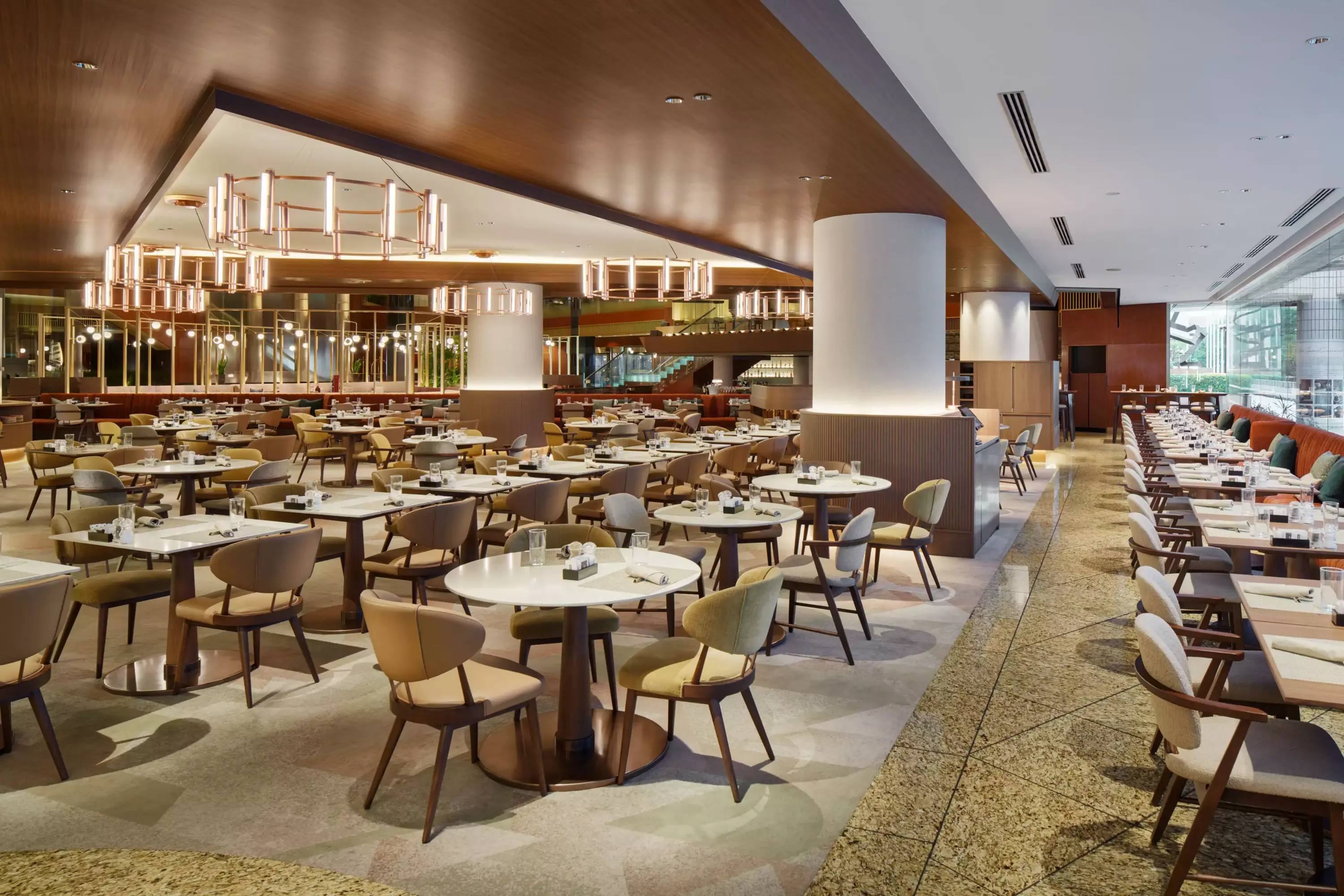 Modern ve ferah Cascade Cafe'nin iç mekanında, ahşap tavan, bakır renkli avizeler ve büyük beyaz sütunlar arasında, beyaz masa tablalı yuvarlak masalar ile bej ve yeşil tonlarında rahat sandalyeler, desenli halı ve fayans zeminde şık bir düzen oluşturuyor.