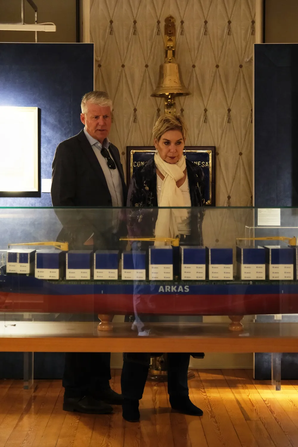 Müze veya sergi ortamında, arkalarında duvarda asılı pirinç bir gemi çanı ve mavi plaket bulunan, önlerinde cam vitrin içinde sergilenen mavi ve kırmızı renkli, üzerinde ARKAS yazan detaylı bir konteyner gemisi modelini dikkatle inceleyen, biri takım elbiseli gri saçlı bir adam ve beyaz şallı bir kadın.