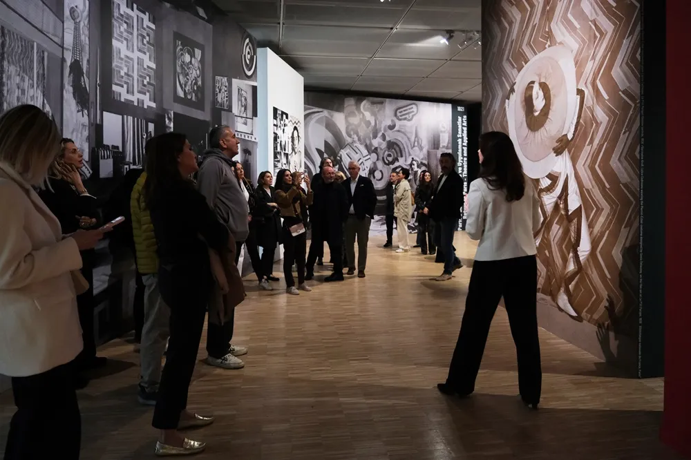 Ahşap zeminli, loş ışıklı bir sanat galerisinde, duvarları siyah-beyaz soyut eserler ve sağda kahverengi tonlarında büyük bir figüratif pano ile kaplı, eserleri ilgiyle inceleyen yaklaşık yirmi kişilik bir ziyaretçi grubu, bu kültürel sanat sergisinin canlı atmosferini doğal bir şekilde yansıtıyor.