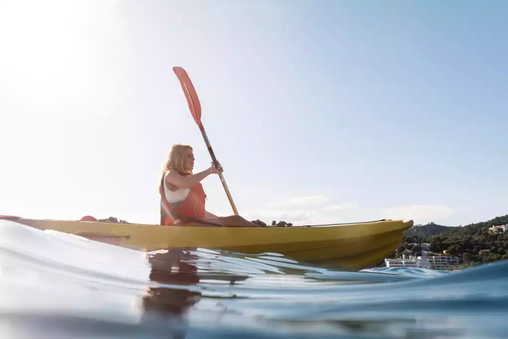 Dalyan'da kayak yapmanın keyfini süren, turuncu can yeleği giymiş sarışın bir kadın, sarı kanosuyla parlayan güneşin altında sakin sular üzerinde ilerlerken, alçak açılı fotoğrafta ön planda hafif dalgalı su yüzeyi, arka planda ise yeşil tepeler ve kıyıdaki binalar dikkat çekiyor.