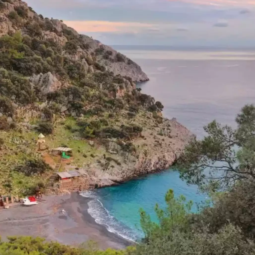 Dalyan'da, kayalık ve yeşilliklerle kaplı yamaçlar arasında kalan, masmavi berrak denizi ve koyu renkli kumsalıyla dikkat çeken bu tenha koyda, sahilde birkaç küçük kulübe ile beyaz bir tekne yer alıyor; alacakaranlığın pembeleşen gökyüzü altında huzurlu bir doğal manzara sunuyor.