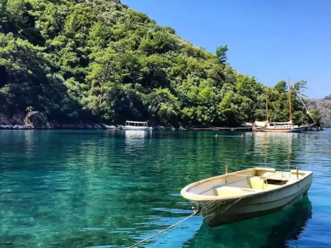 Dalyan bölgesindeki berrak turkuaz renkli sularda demirli küçük, bej renkli bir kayık ve yemyeşil ağaçlarla kaplı tepelerin çevrelediği sakin koyda, uzakta duran diğer teknelerle birlikte huzurlu bir doğal güzellik manzarası.