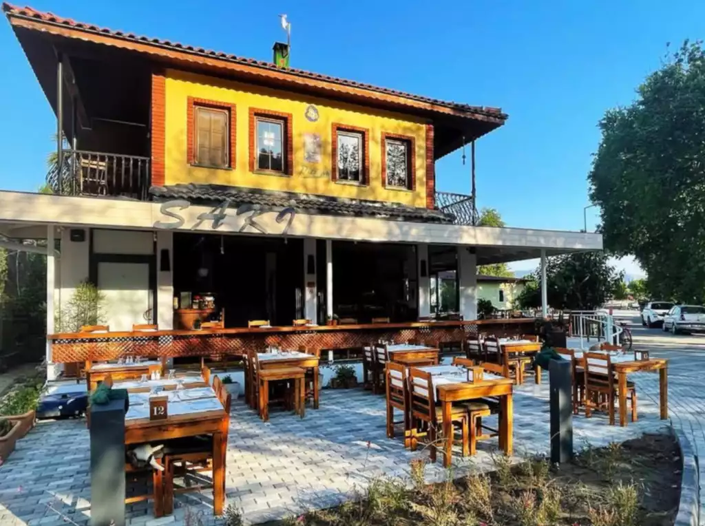 Dalyan'da bulunan, sarı renkli cephesi ve ahşap masaların sıralandığı taş döşemeli geniş açık hava oturma alanıyla sıcak bir atmosfer sunan Saka restoranı, pırıl pırıl bir yaz gününde misafirlerini ağırlamaya hazır görünüyor.