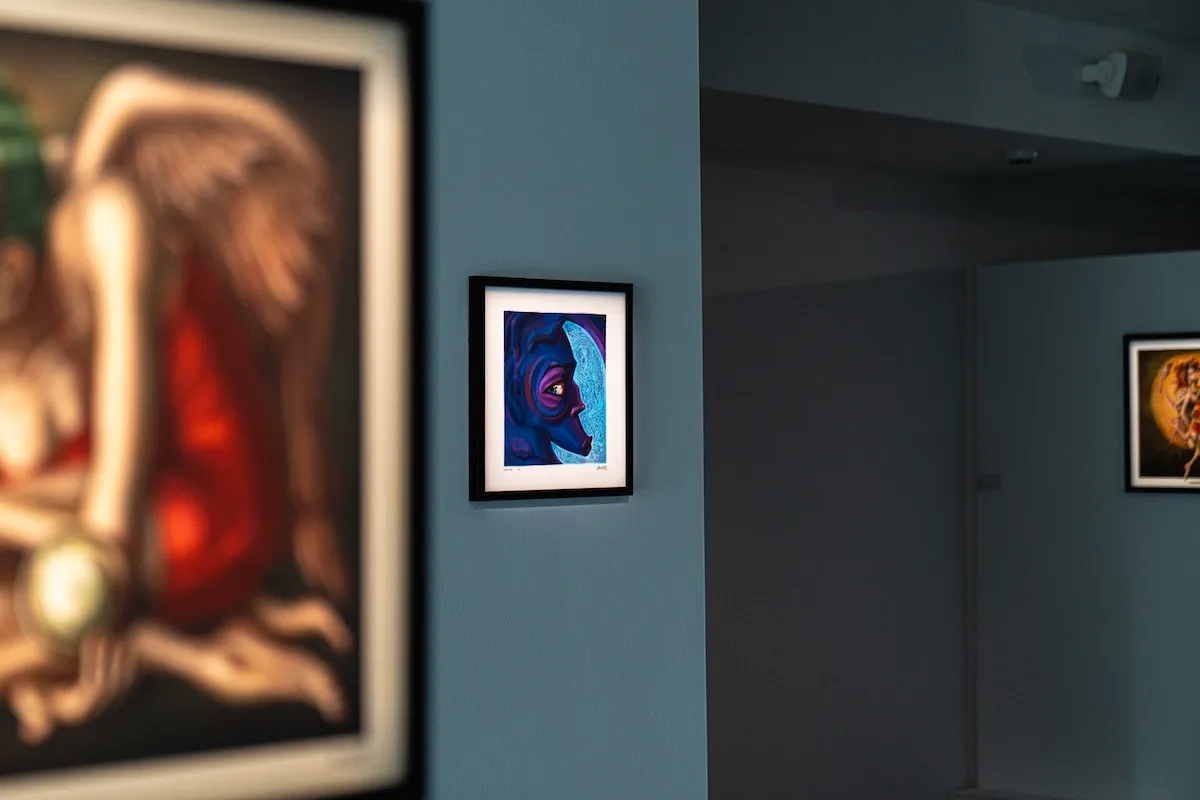 Decollage Art Space'te sergilenen, mavi ve mor tonlarında stilize bir yüzü betimleyen çerçeveli modern bir tablo, soluk mavi bir duvarda odak noktası olarak öne çıkarken, flu görünen başka tablolar sanat galerisi atmosferini tamamlıyor.