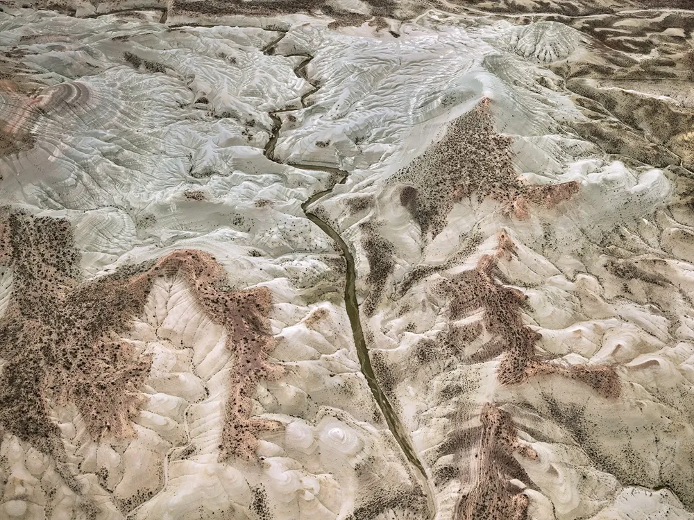 Edward Burtynsky'nin Dönüşen Yeryüzü sergisinden, havadan çekilmiş bu geniş fotoğraf, erozyonla derinlemesine şekillenmiş, kıvrımlı açık renkli tepeler ve vadiler arasında akan dar bir su yolu ile seyrek bitki örtüsünün yayıldığı, insan etkisiyle dönüşen doğal bir manzarayı gözler önüne seriyor.