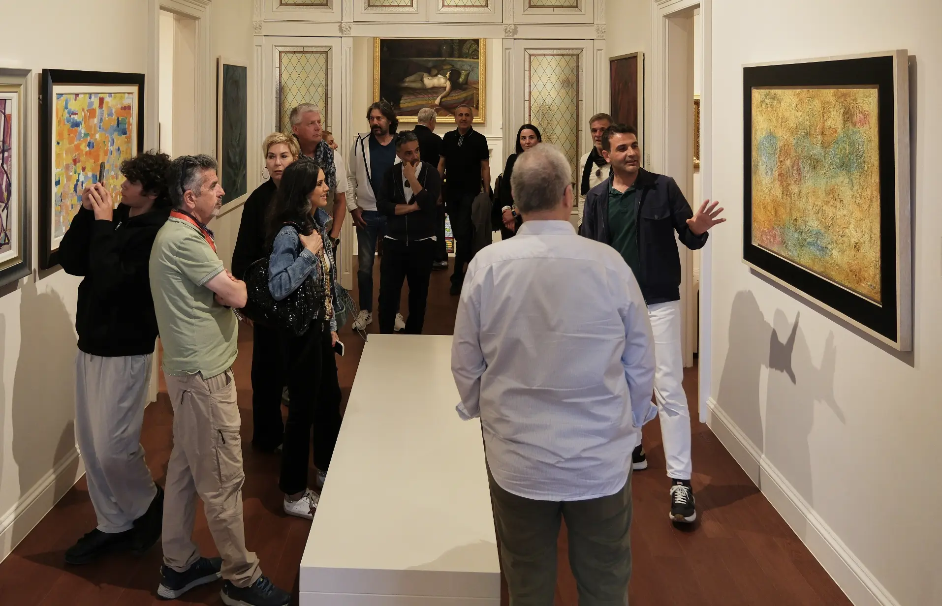 Bir sanat galerisinde düzenlenen sergide, bir rehberin eşliğinde aralarında gençlerin de bulunduğu çeşitli ziyaretçiler, duvarlardaki soyut ve modern sanat eserlerini dikkatle inceliyor, sağdaki rehber sarı ve yeşil tonlardaki tabloyu açıklarken bir genç soldaki eseri fotoğraflıyor.