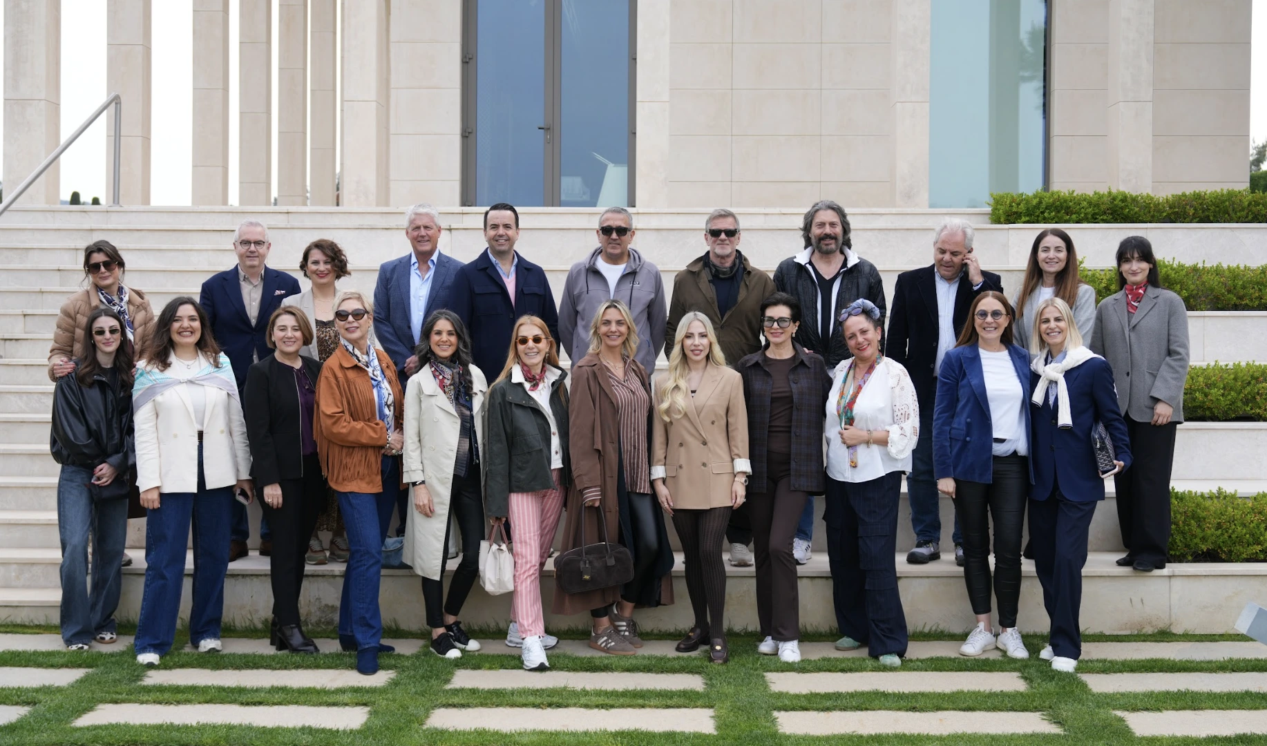 Çeşitli yaş gruplarından yaklaşık 26 erkek ve kadından oluşan bir grup, geniş pencereli modern bir binanın önündeki açık alanda, basamaklarda ve çimenli zeminde şık iş ve günlük kıyafetleriyle sıralanmış, gülümseyerek objektife poz veriyor.