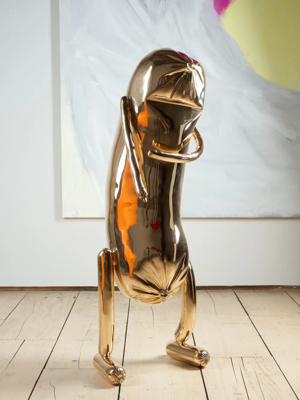 Erwin Wurm'un, parlak ve yansıtıcı altın rengi yüzeye sahip, şişirilmiş balon benzeri formuyla göğsünde kolları kavuşturulmuş, bacakları ve buruşuk baş benzeri üst kısmıyla düşünen bir insan figürünü çağrıştıran modern heykeli, açık renk tahta zeminde, arkasındaki pastel tonlarda soyut tablo önünde duruyor.