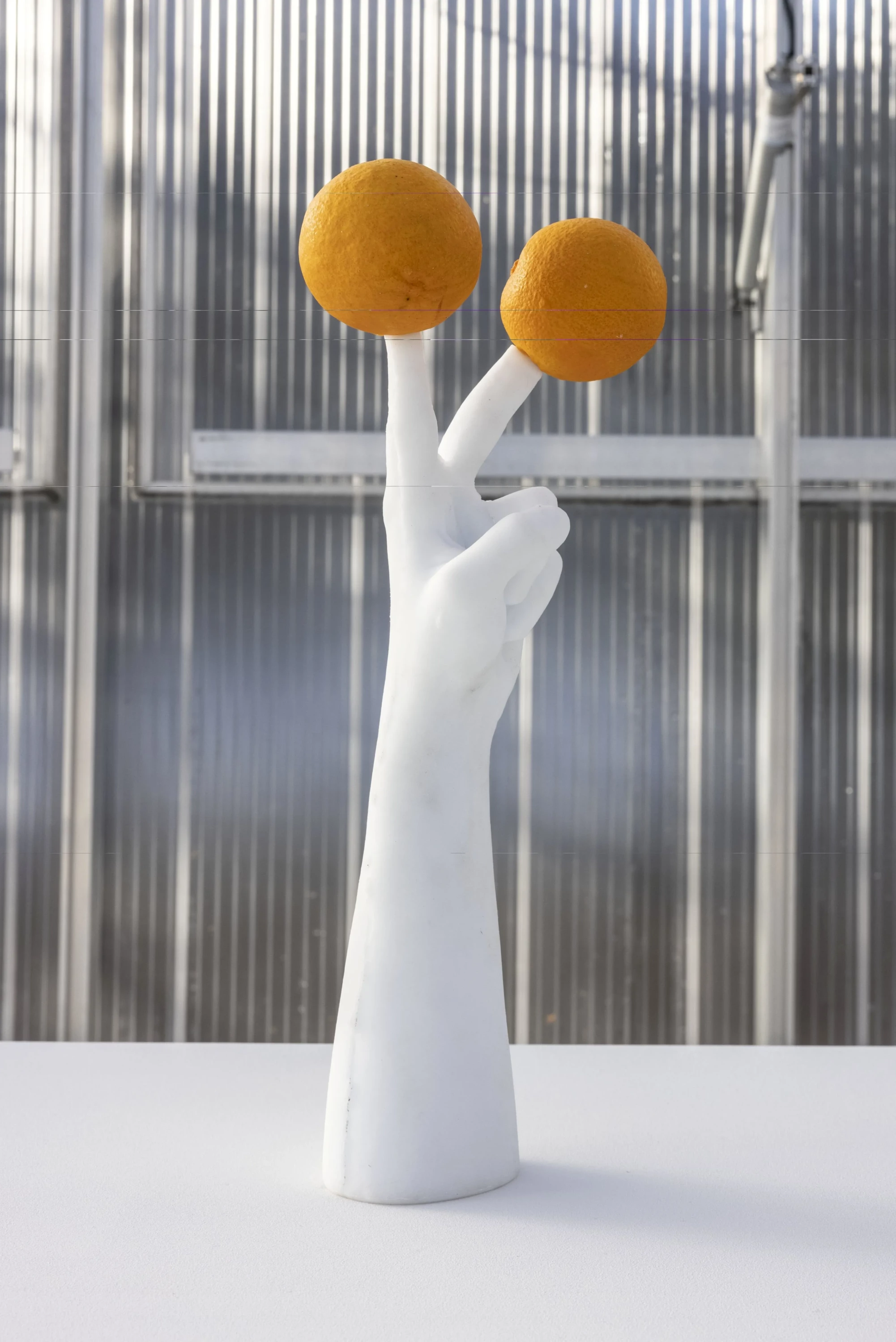 Erwin Wurm'un Murano camından yapılmış, iki parmağı yukarı kalkık, her bir parmağında parlak birer portakal dengelemiş beyaz el heykeli, dikey çizgili gri bir duvarın önündeki beyaz kaide üzerinde, sıra dışı bir portakal ağacı imgesi sunuyor.