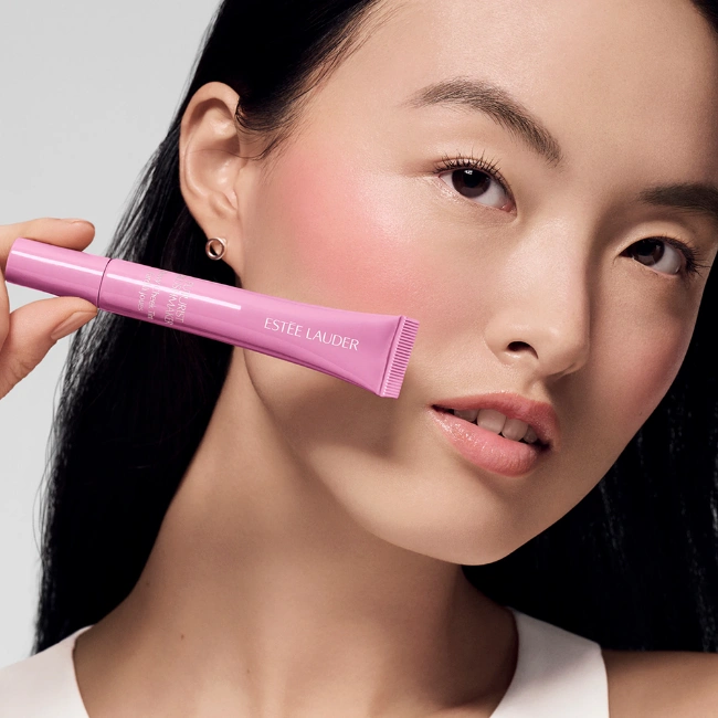 Kameraya doğrudan bakan, yanaklarında doğal, ıslak pembe bir ışıltı taşıyan koyu saçlı model, elinde tuttuğu pembe renkli Estee Lauder Pure Color Envy Dewy Cheek Tint tüpüyle ürünün sunduğu nemli ve canlı yanak görünümünü öne çıkarıyor.