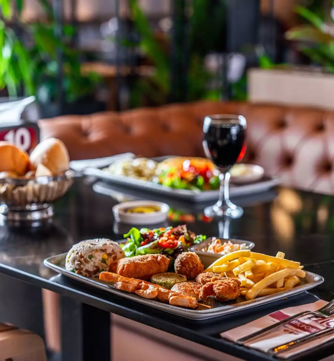 Restoran masasında, çeşitli kızarmış ve ızgara et parçaları, altın rengi patates kızartması, taneli pilav, yeşil salata ile birlikte sunulan zengin bir ana yemek tabağı ön planda dururken, bulanık arka planda kahverengi deri koltuklar ve yeşil bitkilerle donatılmış rahat bir ortamda bir kadeh kırmızı şarap ve ekmek sepeti görülüyor.