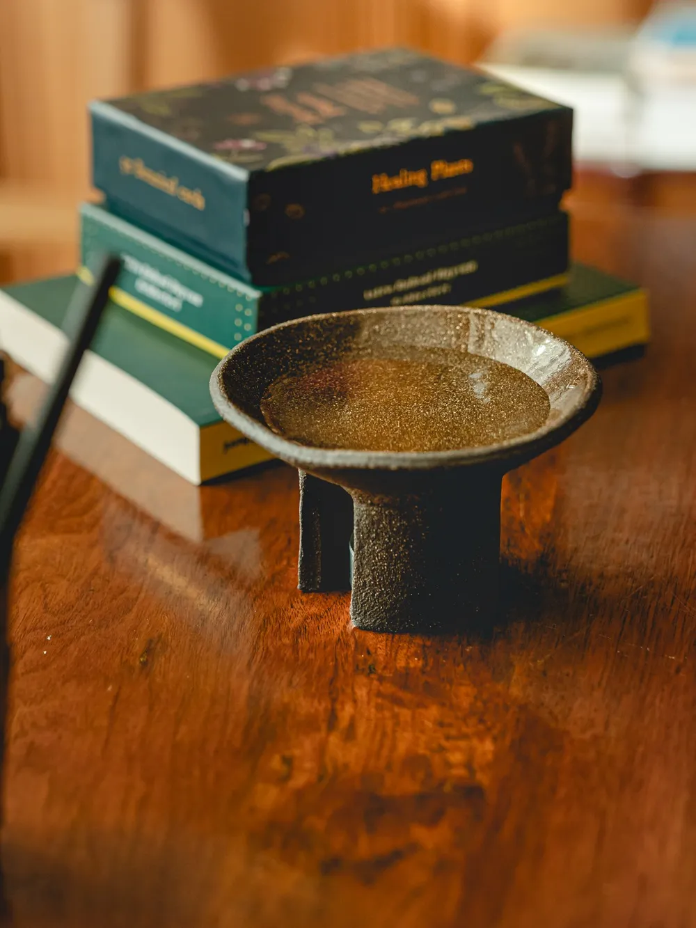 Koyu renkli, dokulu, ayaklı bir seramik kapta parıldayan altın rengi sıvı veya ince taneli karışımın sıcak tonlu ahşap bir yüzeyde durduğu, arka planda Healing Plants yazılı kutular ve kitapların seçildiği bu görsel, Feng Shui ve Aromaterapi uygulamalarına gönderme yaparak dingin bir ambiyans oluşturuyor.
