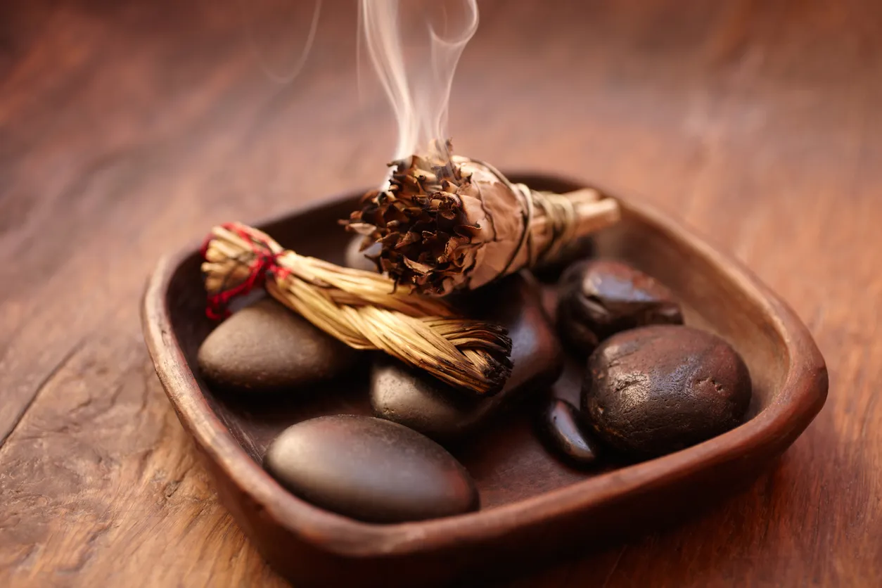 Feng Shui ve aromaterapi uygulamalarında sıkça görülen, ahşap bir tabakta duran koyu renkli taşlar üzerinde dumanı tüten adaçayı demeti ve yanındaki saz demeti, ortam arındırma ve enerji dengeleme ritüellerini çağrıştırıyor.