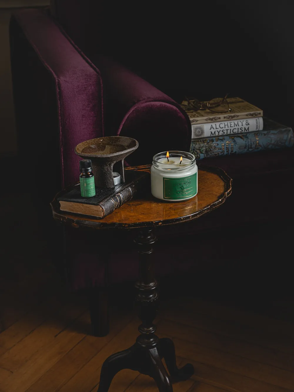 Loş bir odada, mor kadife bir koltuğun yanında duran oymalı antika bir sehpanın üzerinde, huzur verici bir atmosfer oluşturan iki fitilli yanan bir aromaterapi mumu, esansiyel yağ şişesi ve koyu renkli bir buhurdanlık görülüyor; bu düzenleme Feng Shui ve ruhsal dinginlik arayışını yansıtıyor.