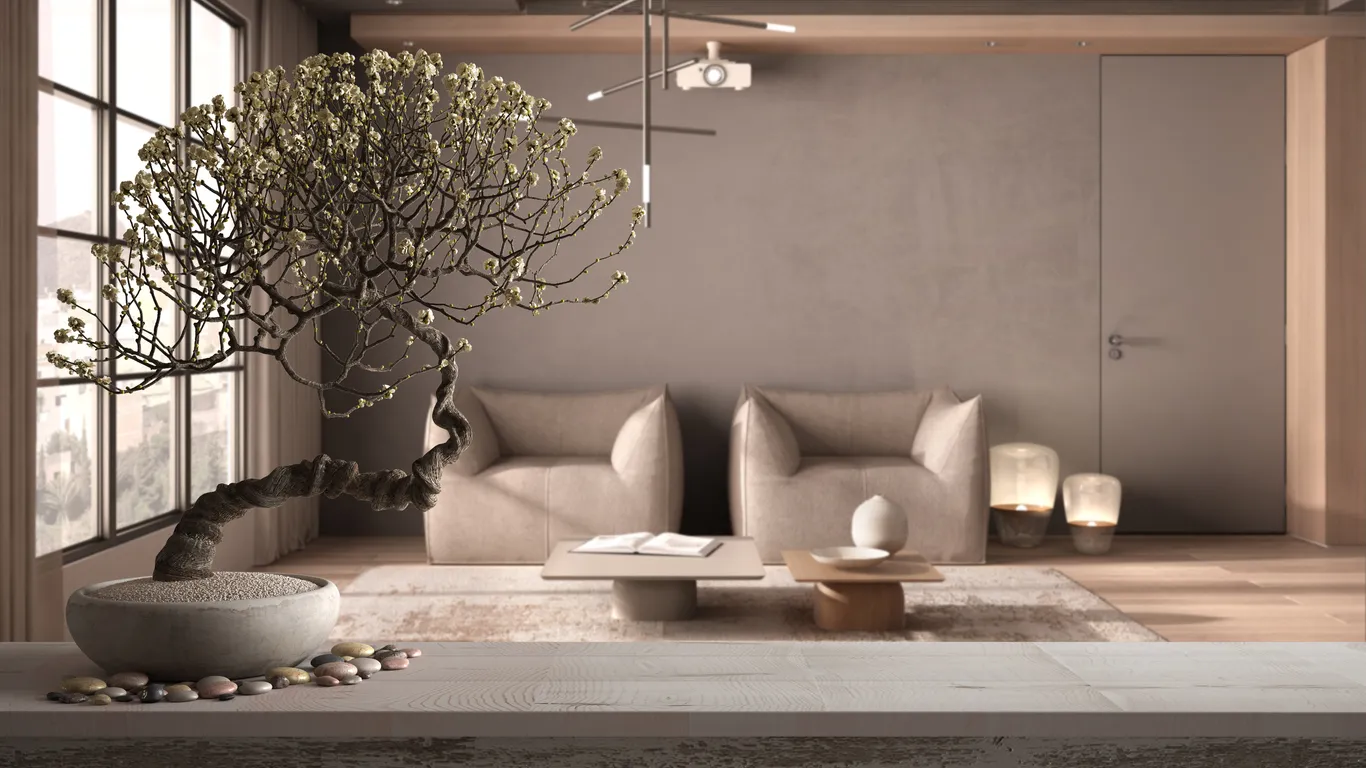 Ön planda, zarif, kıvrımlı gövdeli ve açık renkli çiçek açmış dalları olan bir bonsai ağacı, minimalist bir saksıda, pürüzsüz taşlarla birlikte beyaz ahşap bir yüzeyde dururken, arka planda bej renkli iki koltuğun, modern sehpa ve lambaların bulunduğu sakin, minimalist bir oturma odası manzarası, iç mekanda denge ve uyum sağlayan Feng Shui ilkelerini yansıtıyor.