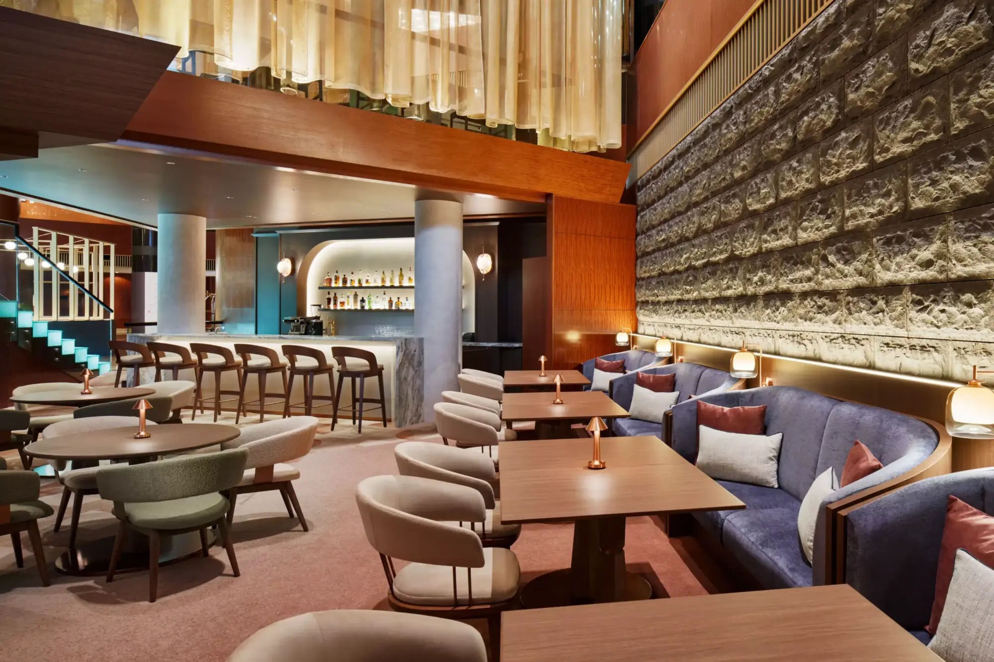 Atrium Lounge içindeki Genever Lobby Bar, sağda rahat mavi kadife banketler ve dokulu taş duvar, solda arkası ışıklı şişelerle dolu mermer bar ve modern bar tabureleri, ortada ise farklı tasarımlı sandalyeler ve bakır renkli lambalarla süslenmiş ahşap masalarla sıcak bir ortam sunuyor.