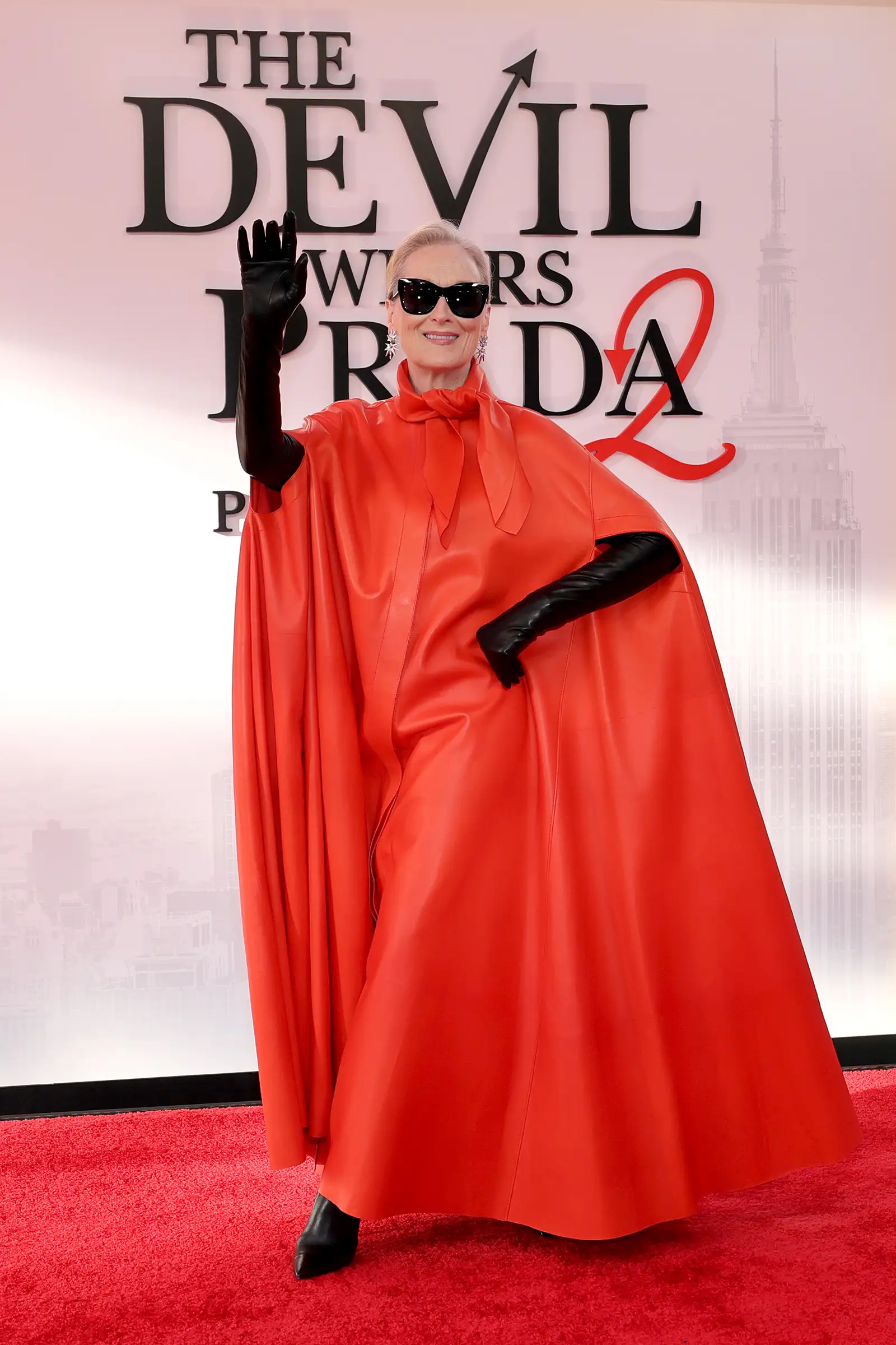 Meryl Streep, ikonik The Devil Wears Prada filminin devamı temalı bir etkinlikte, göz alıcı parlak kırmızı pelerin elbisesi, siyah uzun eldivenleri ve güneş gözlükleriyle kırmızı halıda neşeyle el sallayarak poz veriyor, arkasında THE DEVIL WEARS PRADA 2 yazısı ve New York silüeti seçiliyor.