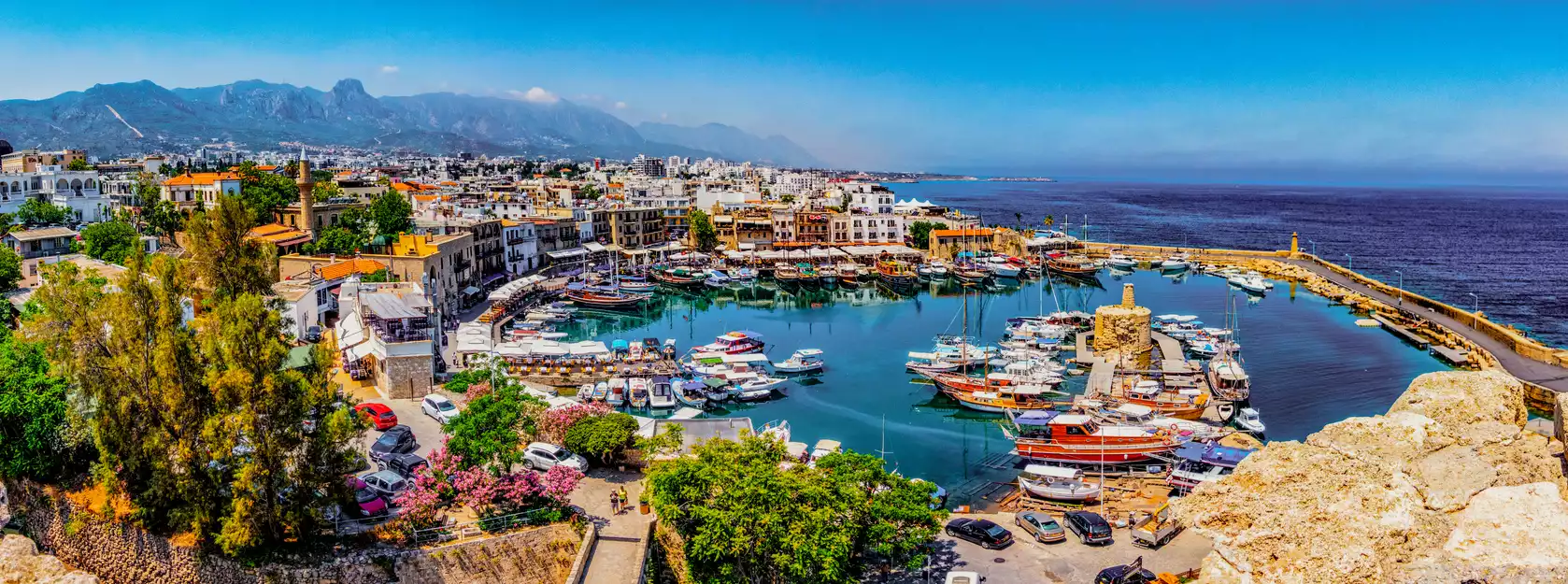 Akdeniz'in berrak sularında demirlemiş sayısız tekne ve yatın göz alıcı bir şekilde sıralandığı, masmavi gökyüzü altında parlayan Kıbrıs'ın tarihi limanında canlı bir gün yaşanırken, arka planda yükselen görkemli dağlar ve kıyıya dizilmiş geleneksel evler bu pitoresk manzarayı tamamlıyor.