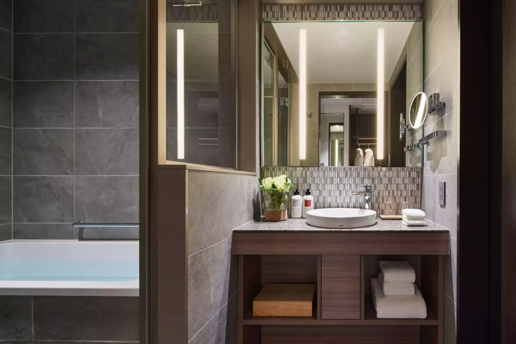 Mizu Suite banyosunda, modern koyu gri fayanslarla döşenmiş bir küvetin yanındaki ahşap tezgah üzerinde, beyaz oval lavabo, dekoratif aynalar, taze çiçekler ve özenle katlanmış havlularla donatılmış, ışıklı bir ayna dikkat çekiyor.