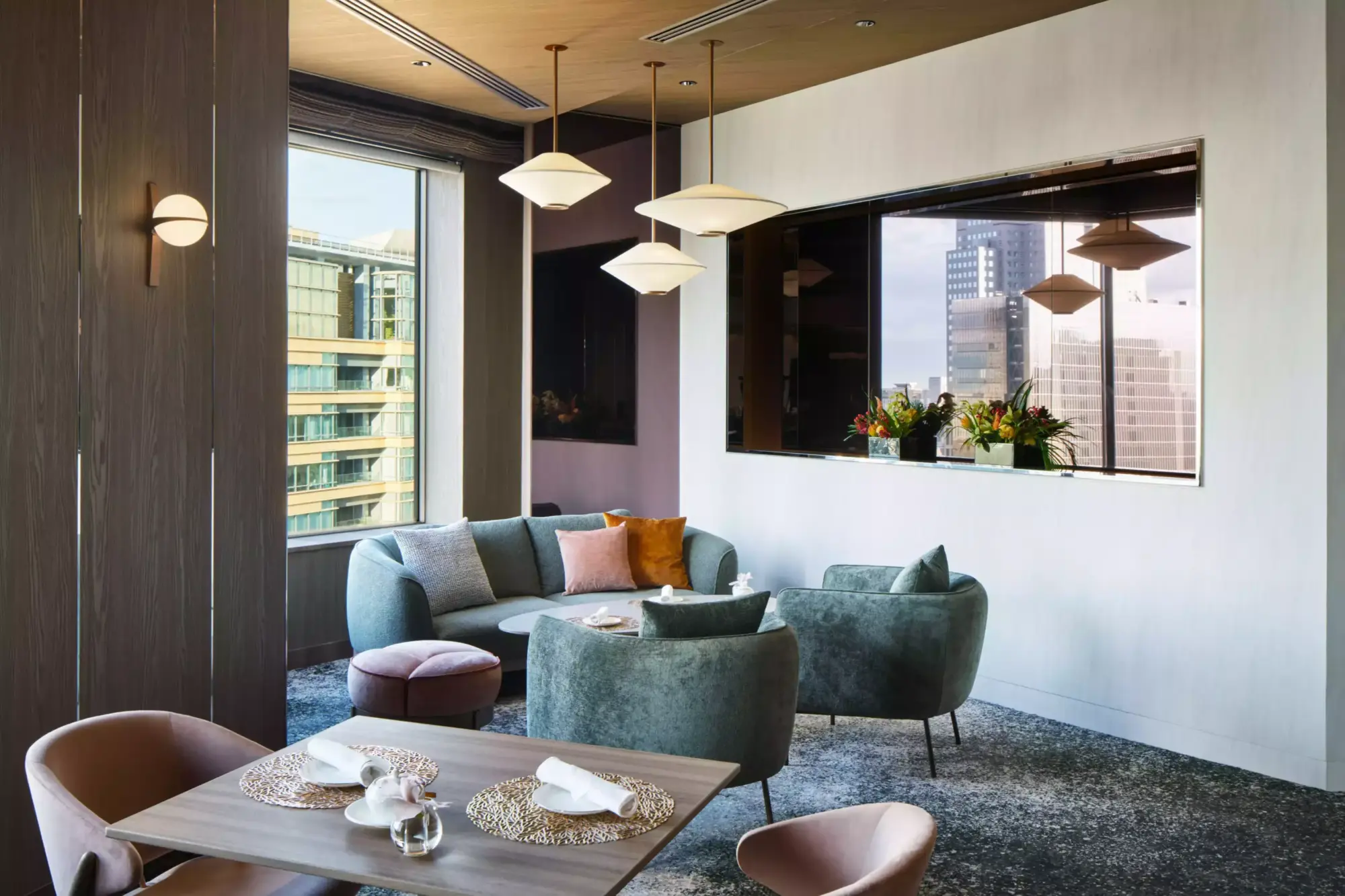 Yeni Club InterContinental Lounge'ın 36. katındaki rahat ve modern oturma alanında, şehir manzaralı büyük camların önünde gri-mavi bir kanepe ve koltuklar, ahşap yemek masası, konik avizeler ve çiçekler lüks bir atmosfer oluşturuyor.