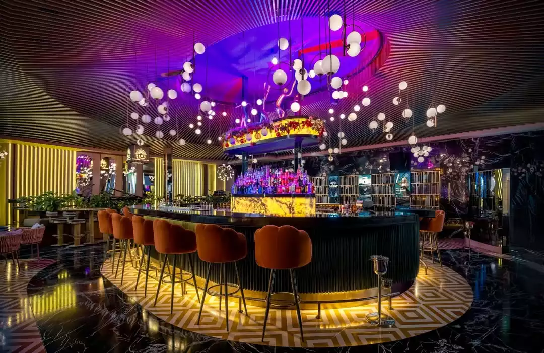 Modern ve lüks bir bar iç mekanı; tavandan sarkan çok sayıda küresel lambanın mor ve mavi tonlarında ambiyans ışıklarıyla aydınlattığı, parlak renkli şişelerle dolu kavisli tezgahının önünde turuncu bar tabureleri sıralanıyor.