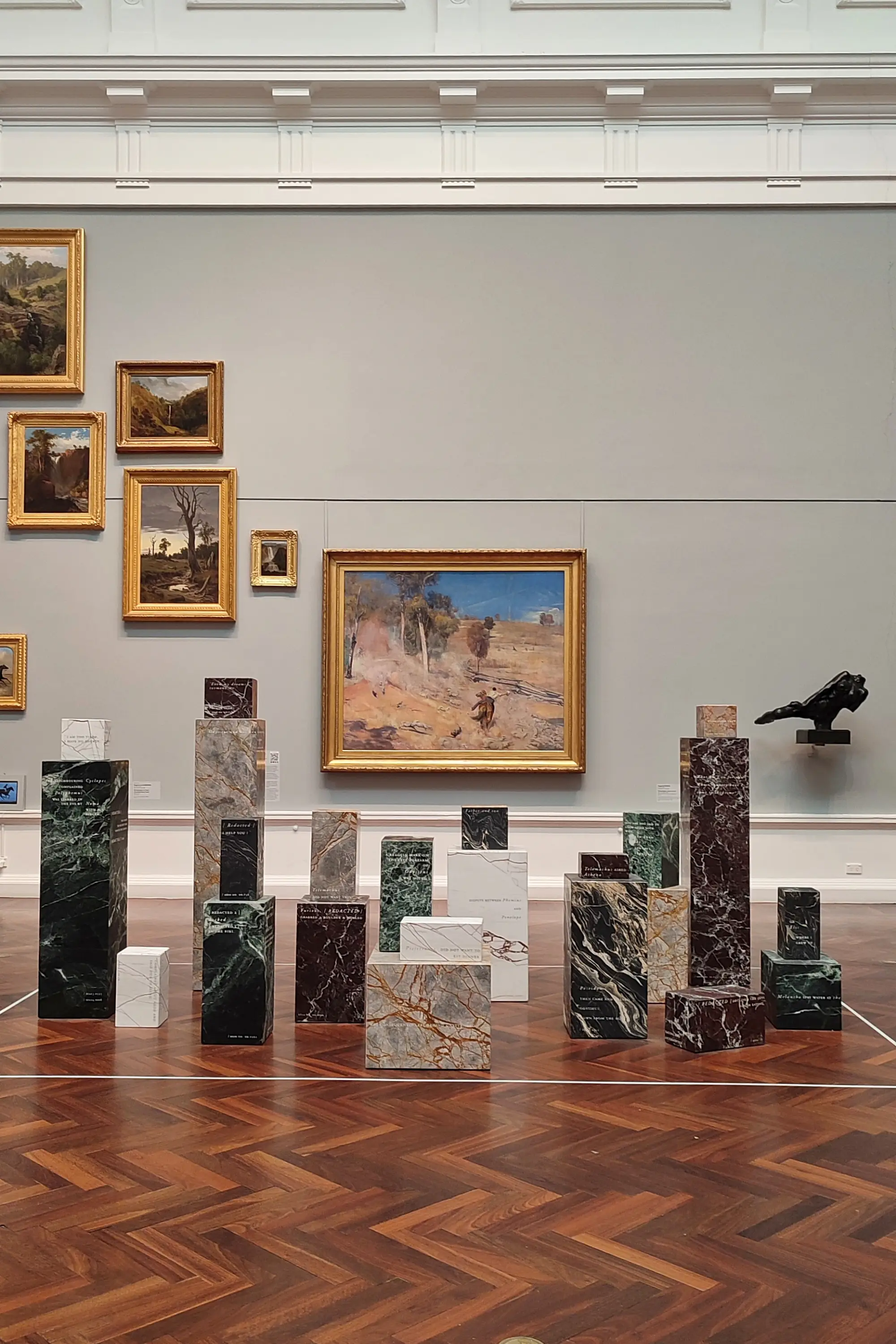 Sanat galerisinde, ahşap parke zemin üzerinde yükselen, üzerlerinde yazılar bulunan çeşitli renk ve desenlerdeki mermer bloklardan oluşan ilgi çekici bir modern sanat enstalasyonu, arkada duvarda asılı geleneksel yağlıboya manzara resimleriyle beraber sergileniyor.
