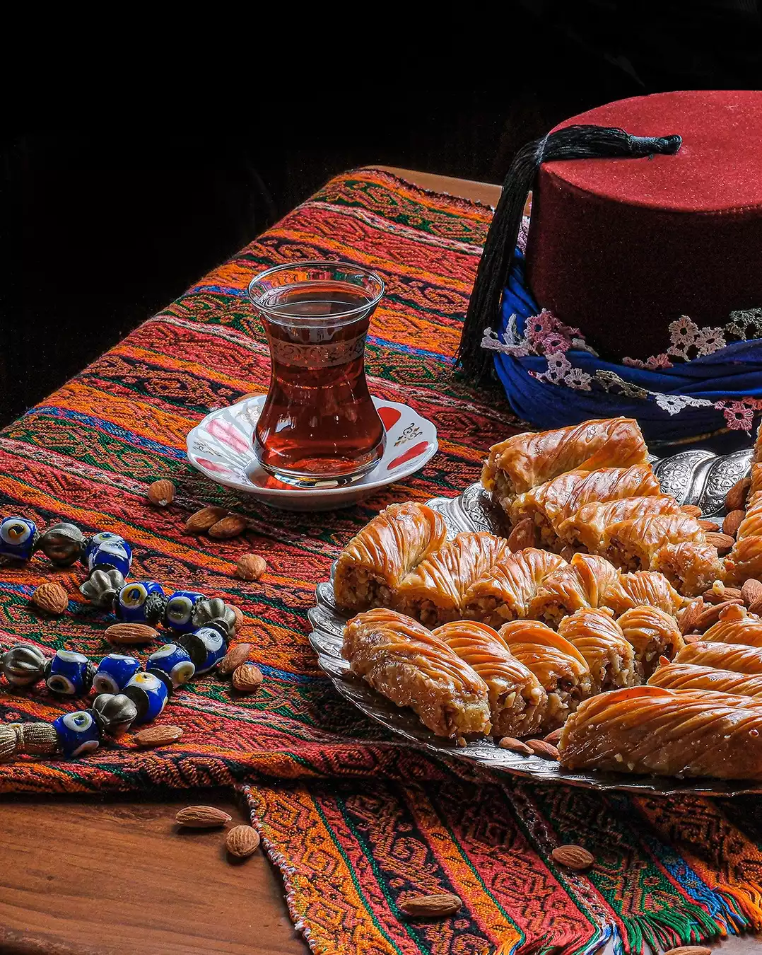 Renkli desenli bir kilimin üzerinde yer alan, cevizli baklavaların dizildiği parlak gümüş bir tepsi, zarif ince belli çay bardağında demli çay, kırmızı fes ve nazar boncuklarının bademlerle süslediği, geleneksel Türk misafirperverliğini yansıtan zengin bir görsel şölen.