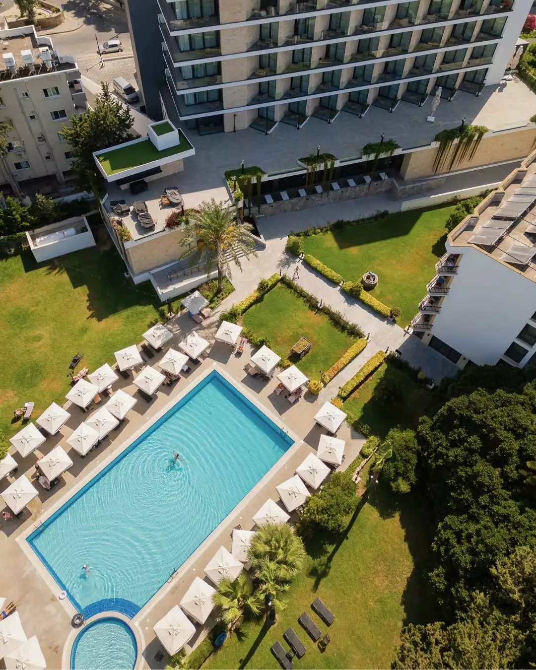 Geniş bir alana yayılmış Pia Bella Otel'in modern mimarili binası, çevresinde yer alan berrak mavi yüzme havuzu, beyaz güneş şemsiyeleri ve şezlonglarla dolu dinlenme alanları ile yemyeşil bahçelerin kuşbakışı görünümü, tesisin davetkar atmosferini sergiliyor.
