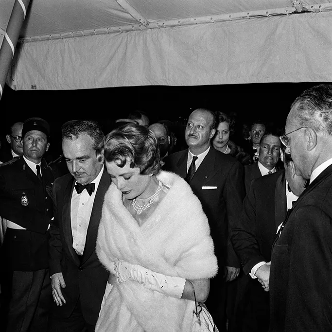 Monako Prensi III. Rainier ve Prenses Grace Kelly, 1958 yılında katıldıkları bir etkinlikte, Grace Kelly'nin başı eğik beyaz kürk şal ve inci kolye takarak yürüdüğü, Rainier'in ise smokinle yanında durduğu anı gösteren siyah beyaz bir fotoğraf.