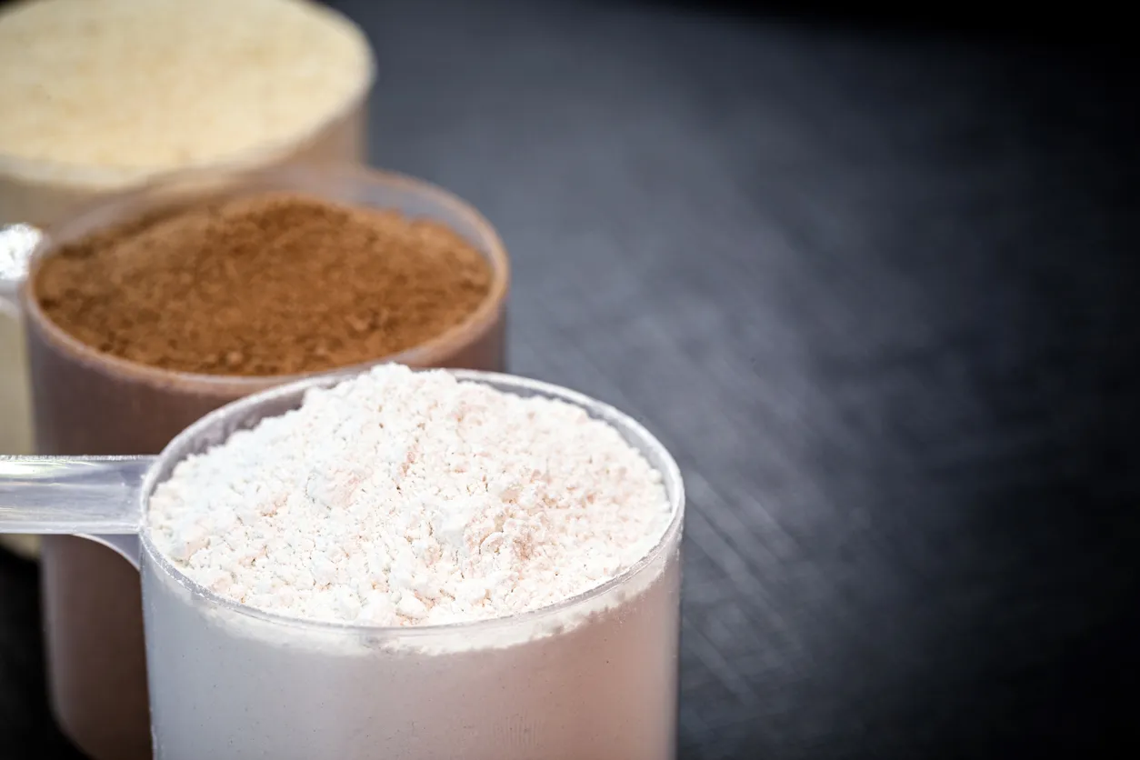 Üç adet şeffaf ölçü kaşığında, en önde açık renkli, ortada kahverengi ve arkada krem rengi olmak üzere farklı türlerdeki protein tozları, protein tozu seçimi yaparken dikkate alınması gereken çeşitli seçenekleri vurgulayacak şekilde koyu bir zemin üzerinde kademeli olarak sıralanmış.