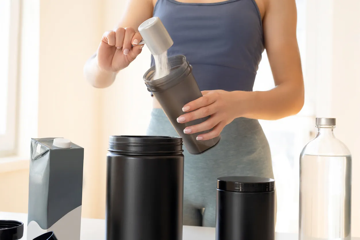 Spor kıyafetli bir kadın, büyük bir kutudan kepçe yardımıyla beyaz protein tozunu koyu gri bir shaker şişesine aktarırken görülüyor; masada ayrıca bir süt kartonu, su şişesi ve başka protein tozu kapları bulunuyor.