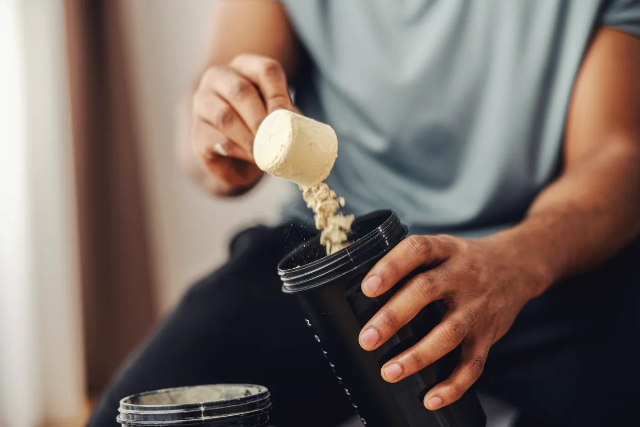 Açık renk tişörtlü bir kişi, bej renkli protein tozunu ölçü kaşığından siyah bir shaker şişesine dikkatle boşaltırken toz parçacıkları havada süzülüyor; bu an, sporcuların beslenme rutinlerinde doğru protein tozu seçerken gösterdiği özeni yansıtıyor.