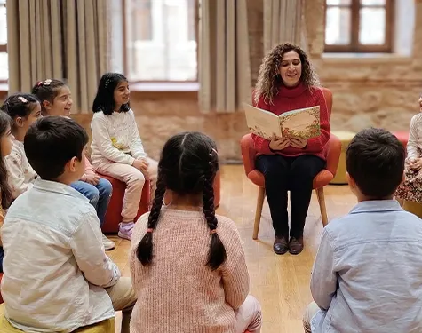 Rami Çocuk ve Sanat Bienali çerçevesinde gerçekleşen interaktif bir atölye etkinliğinde, kırmızı kazaklı ve kıvırcık saçlı bir eğitmen, etrafında dikkatle oturan çocuklara resimli bir kitap okuyarak çocuk edebiyatını deneyimlenebilir sanat alanlarına taşıyor.